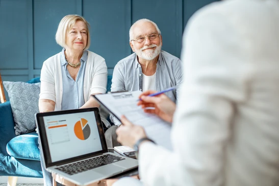 Consultation financière à domicile entre un conseiller et un couple de seniors assis sur canapé bleu