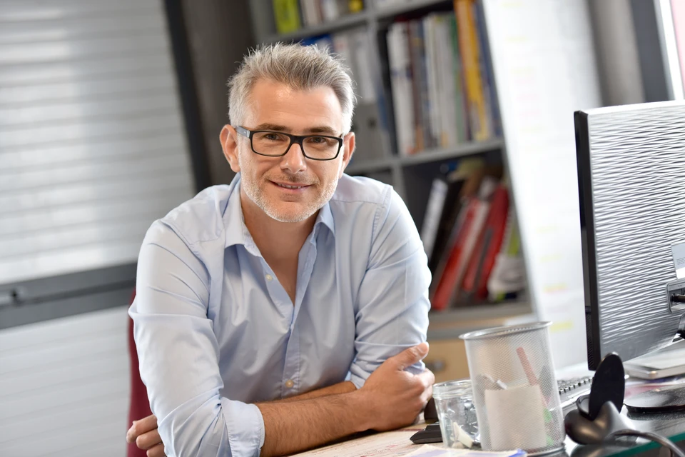 Un homme d'affaires mature avec lunettes et barbe sourit dans un bureau moderne, assis à son poste de travail