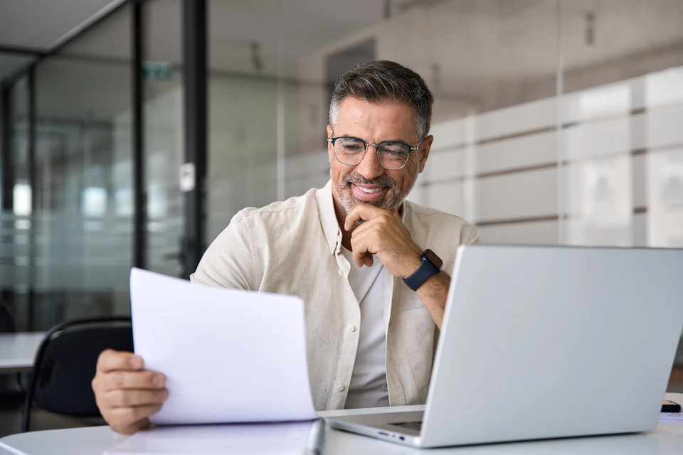 Souriant analyste comptable homme d'affaires latin ou indien tenant des documents, travaillant sur ordinateur portable faisant de la recherche technologique sur le marché du commerce en ligne