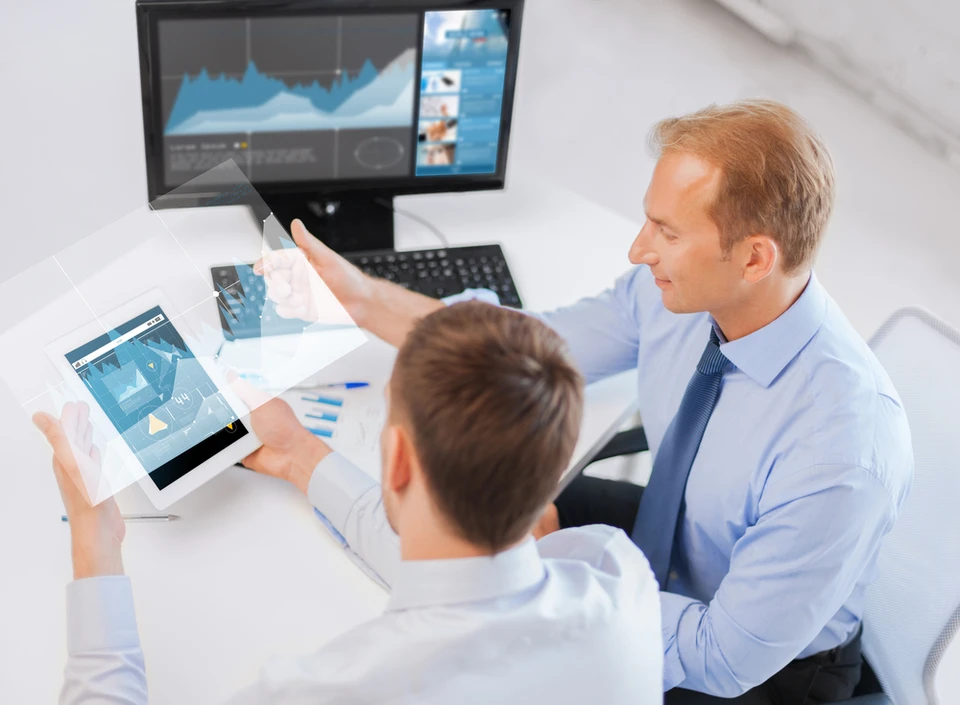 Homme d'affaires en chemise bleue et cravette noire analysant des graphiques et tableaux de données imprimés tout en utilisant un ordinateur portable dans un bureau moderne avec éclairage chaleureux en arrière-plan
